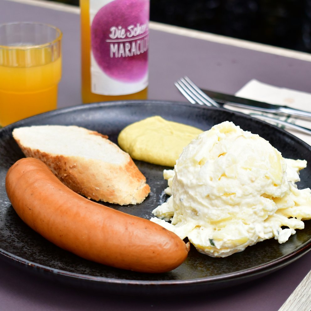 herzhafte-speisen Bockwurst mit Kartoffelsalat und einer Scheibe Baguette auf einem Teller. Daneben steht eine Glasflasche und ein Glas
