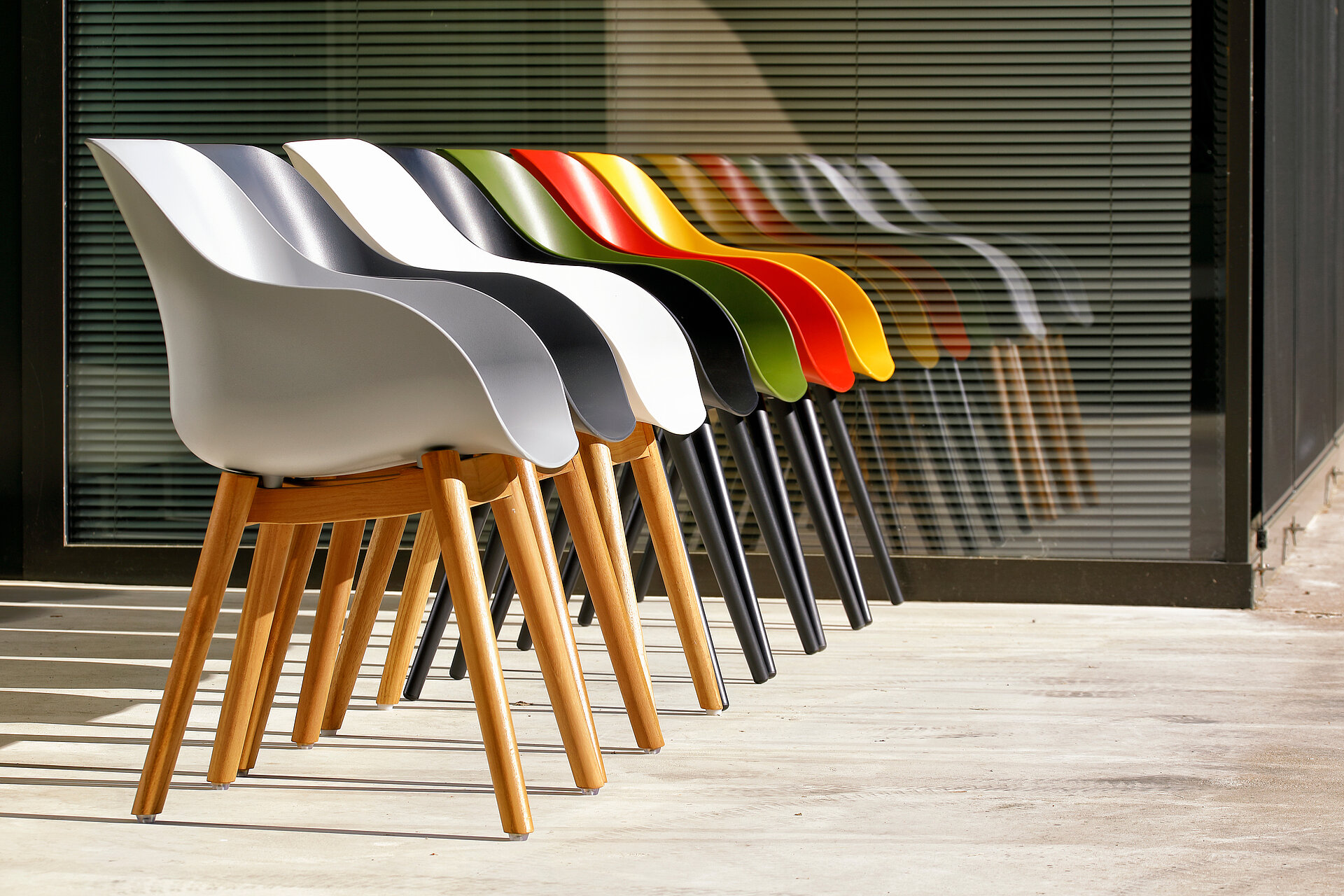 Sieben Stühle der Firma Hartmann, jede Sitzschale hat eine andere Farbe grau, anthrazit, weiß, schwarz grün, rot, gelb. Im Hintergrund sieht man eine Glasscheibe, die von Jalousien verdeckt sind. Bei Dinger’s Gartenmöbel kaufen.