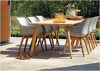 Ein Holztisch auf einer Terrasse daneben sechs Stühle mit Holzbeinen und einer graufarbenen Sitzschale. Auf dem Tisch steht eine Karaffe, drei Gläser, ein Obstkorb und eine Etagere.