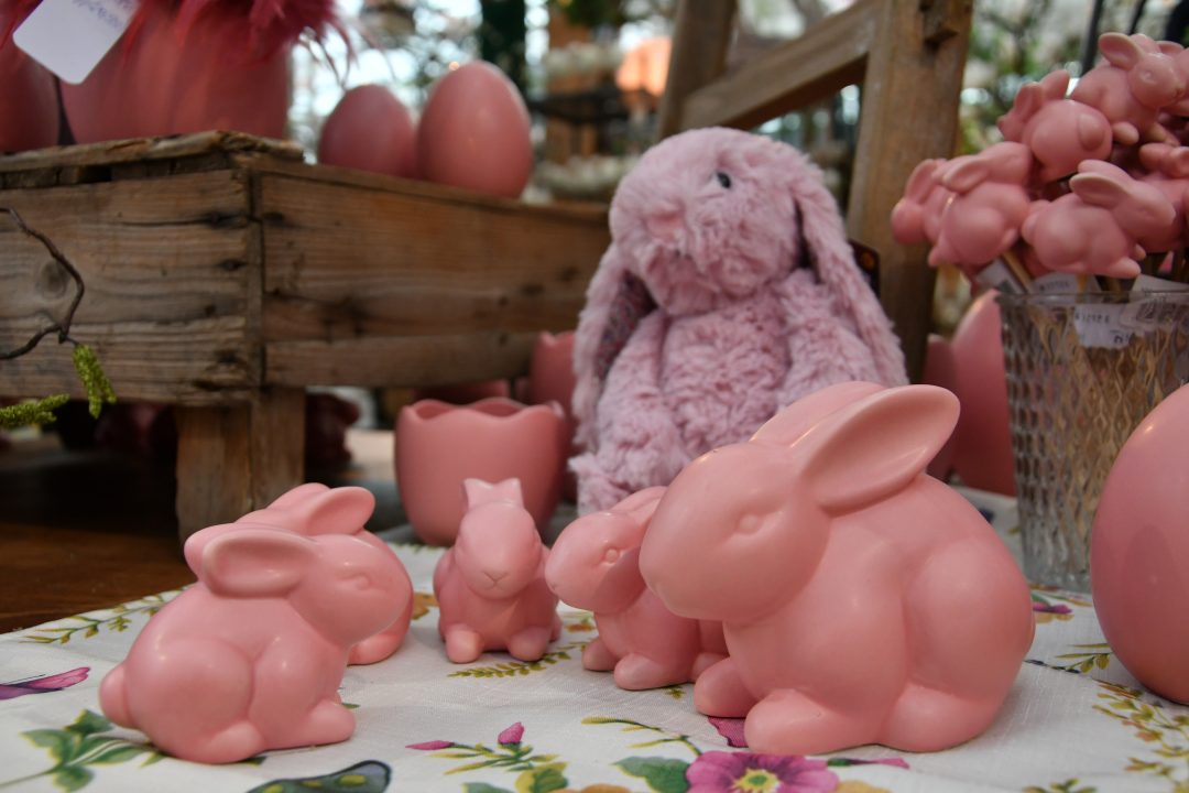 Auf diesem Bild erkennt man rosafarbene Keramik Osterhasen und einen flauschigen Osterhasen in der selben Farbe. Sie stehen auf einer blumigen Tischdecke und im Hintergrund erkennt man weitere österliche Dekoelemente.