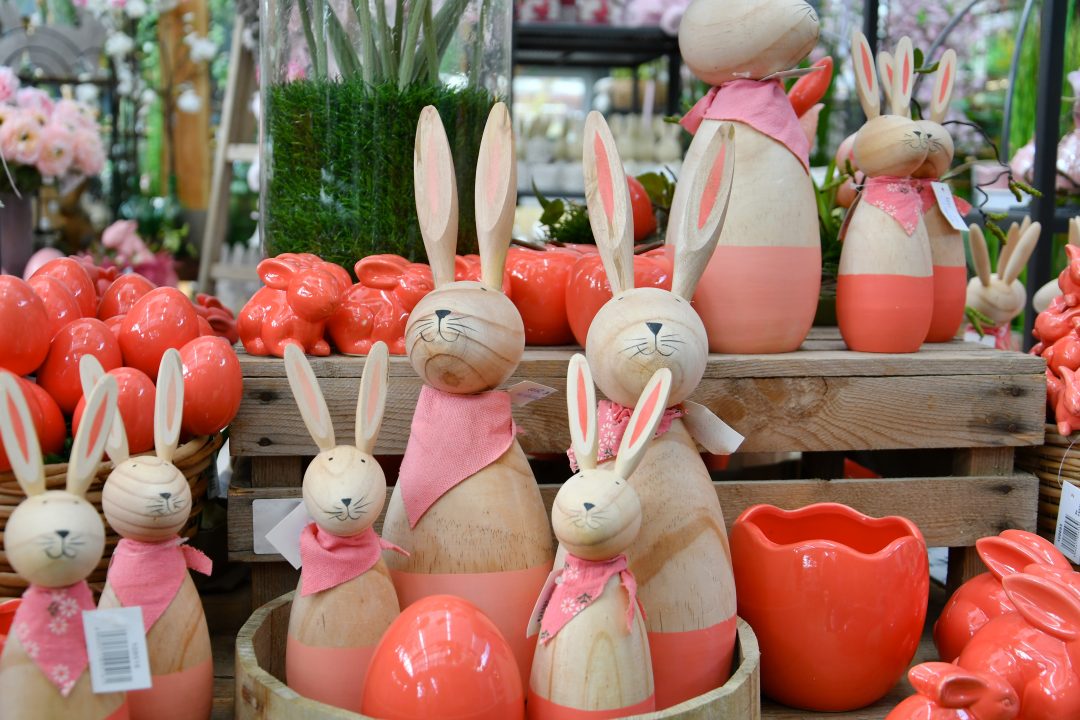 Auf diesem Bild sieht man viele HOlzosterhasen mit rosafarbenen Halstüchern. Daneben stehen diverse Osterhasen und Eier in korallrot, einer Trendfarbe in 2020. Im Hintergrund sind weitere Dekoelemente zu erkennen.