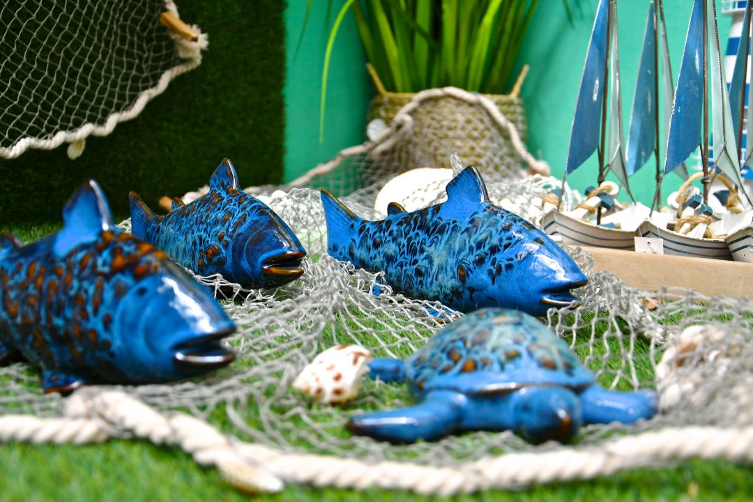 Maritime Dekoration. Blaue Porzellan Fische und eine Schildkröte auf einem Fischernetz. Rechts sieht man kleine blau weiße Segelboote.